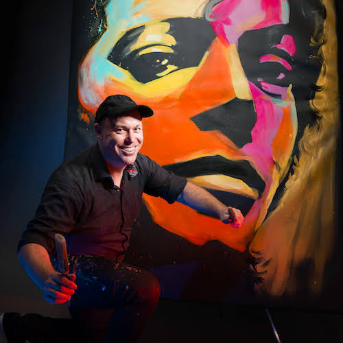 Artist Brad Blaze seated in front of one of his colourful portraits.